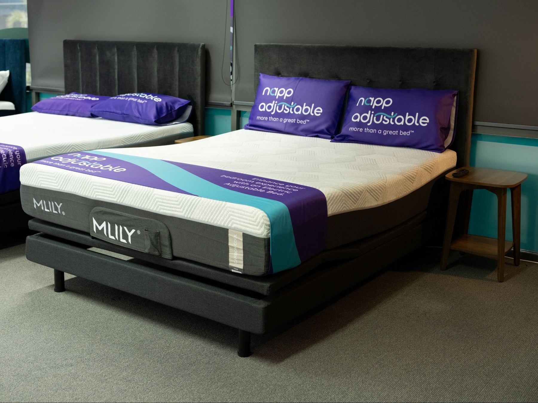 Queen Revitalise adjustable bed shown in a showroom setting, featuring white mattresses with purple and teal bedding, black upholstered headboards, and matching purple pillows.