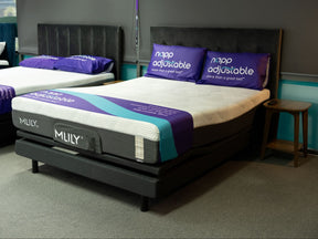 Queen Revitalise adjustable bed shown in a showroom setting, featuring white mattresses with purple and teal bedding, black upholstered headboards, and matching purple pillows.
